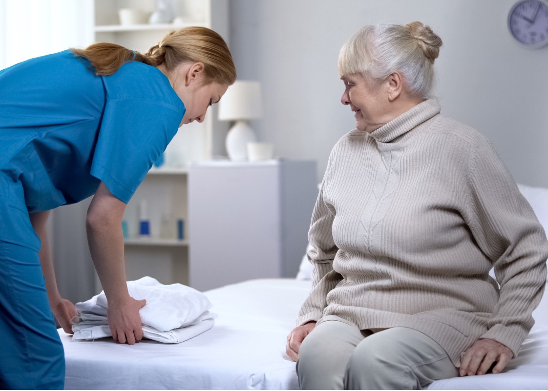 nurse-preparing-clean-bedlinen-to-elderly-female-patient-in-medical-picture-id1150489856